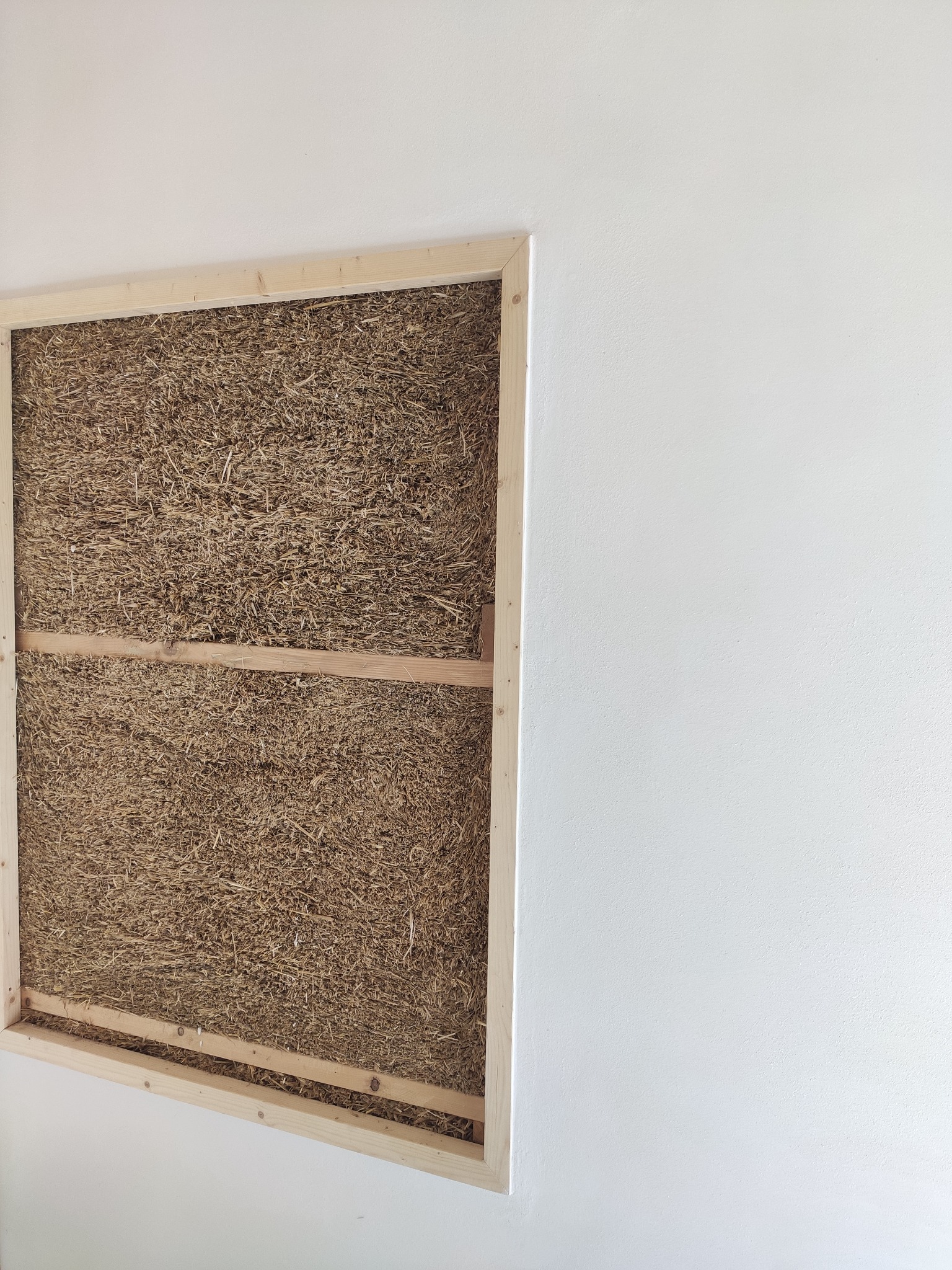 Fragment ściany z wypełnieniem ze słomy w drewnianej ramie, widoczne połączenia elementów ramy, surowa biała ściana obok.
