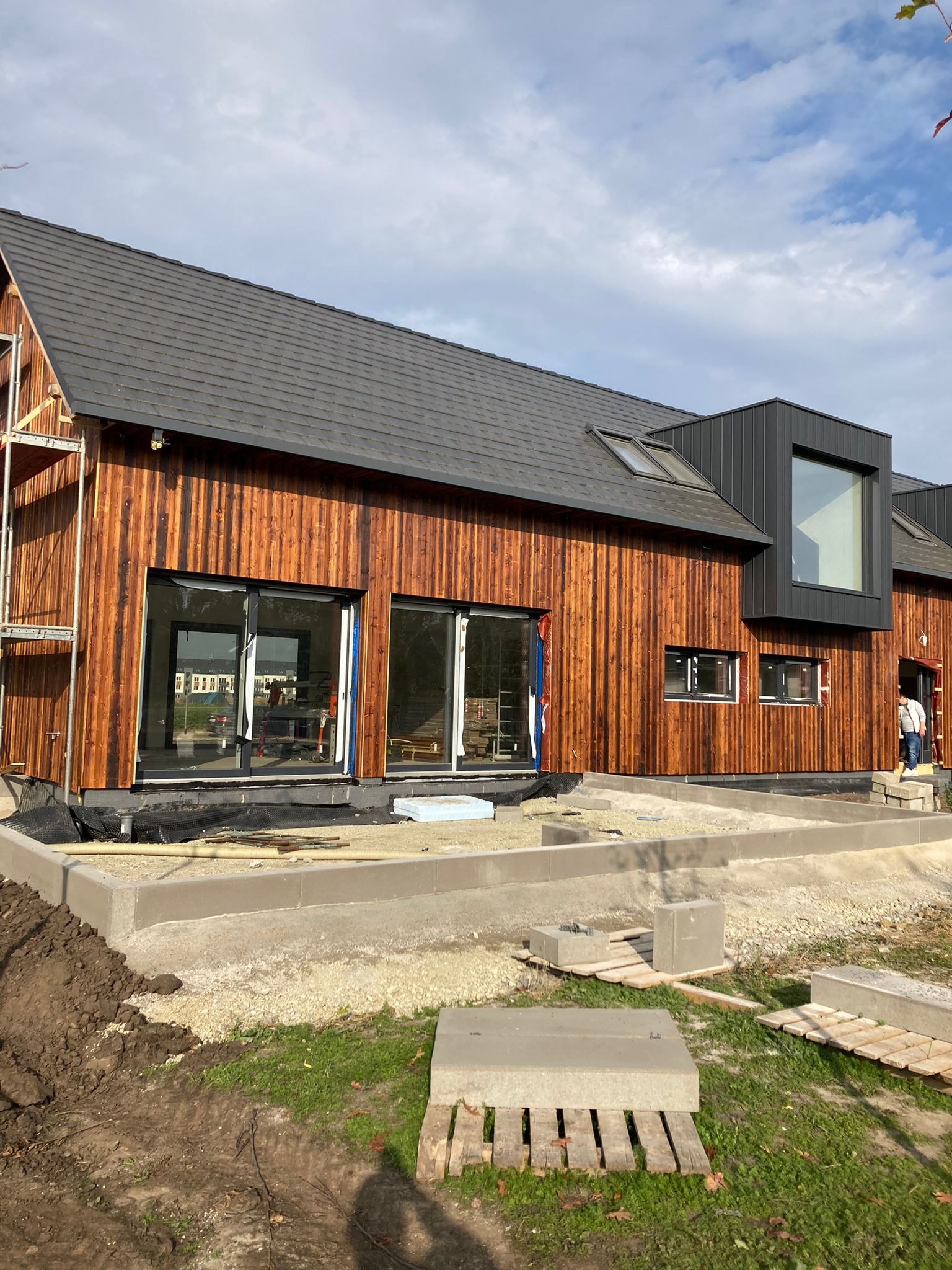 Nowoczesny dom z drewnianą elewacją w trakcie wykończenia, widoczne rusztowania, duże okna przesuwne i betonowy fundament.