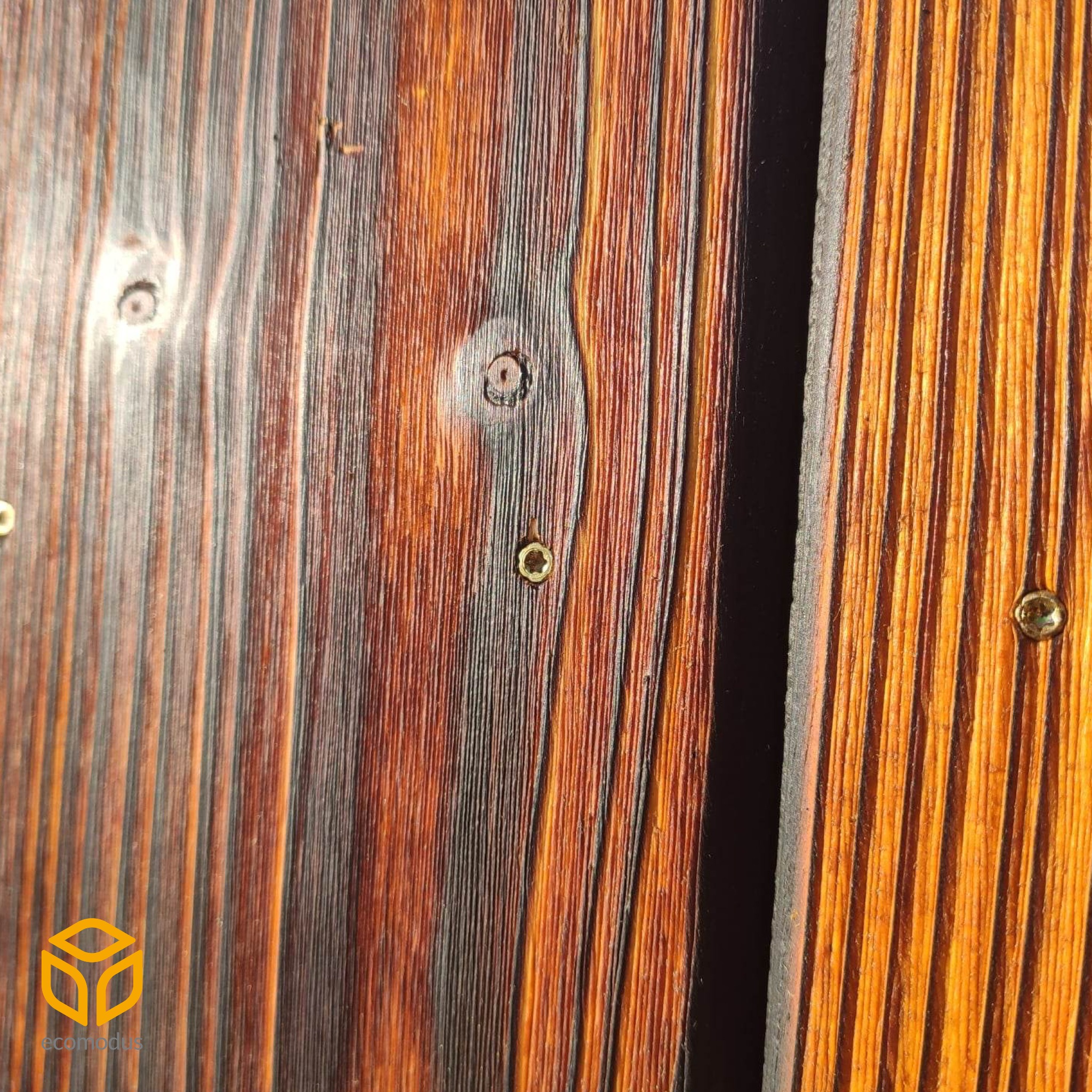 Zbliżenie na fragment elewacji z ciemnego, lakierowanego drewna z widocznymi śrubami mocującymi i logo firmy w lewym dolnym rogu.