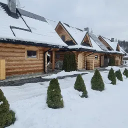 Zewnętrzne jednostki klimatyzacyjne w drewnianych obudowach przy domkach w stylu góralskim, zimowy krajobraz z ośnieżonymi drzewami.