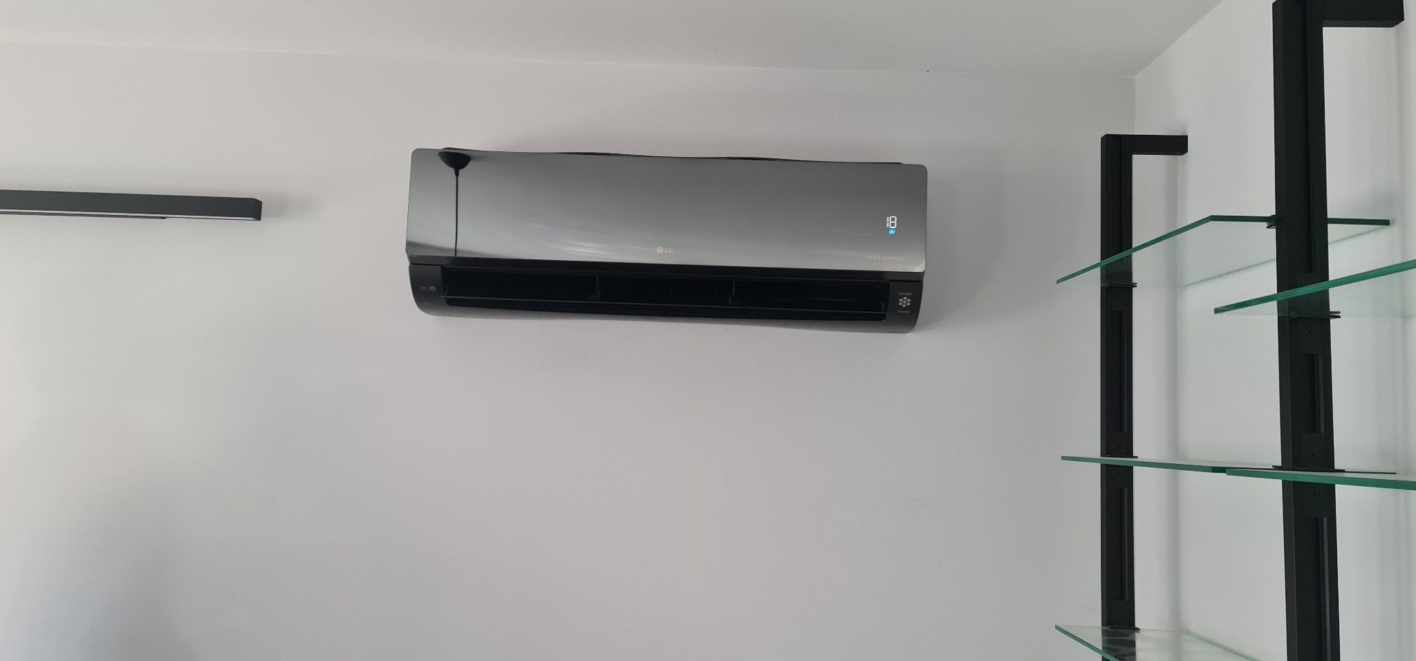 Szary klimatyzator LG typu split zamontowany na białej ścianie, obok czarne półki ze szklanymi blatami.