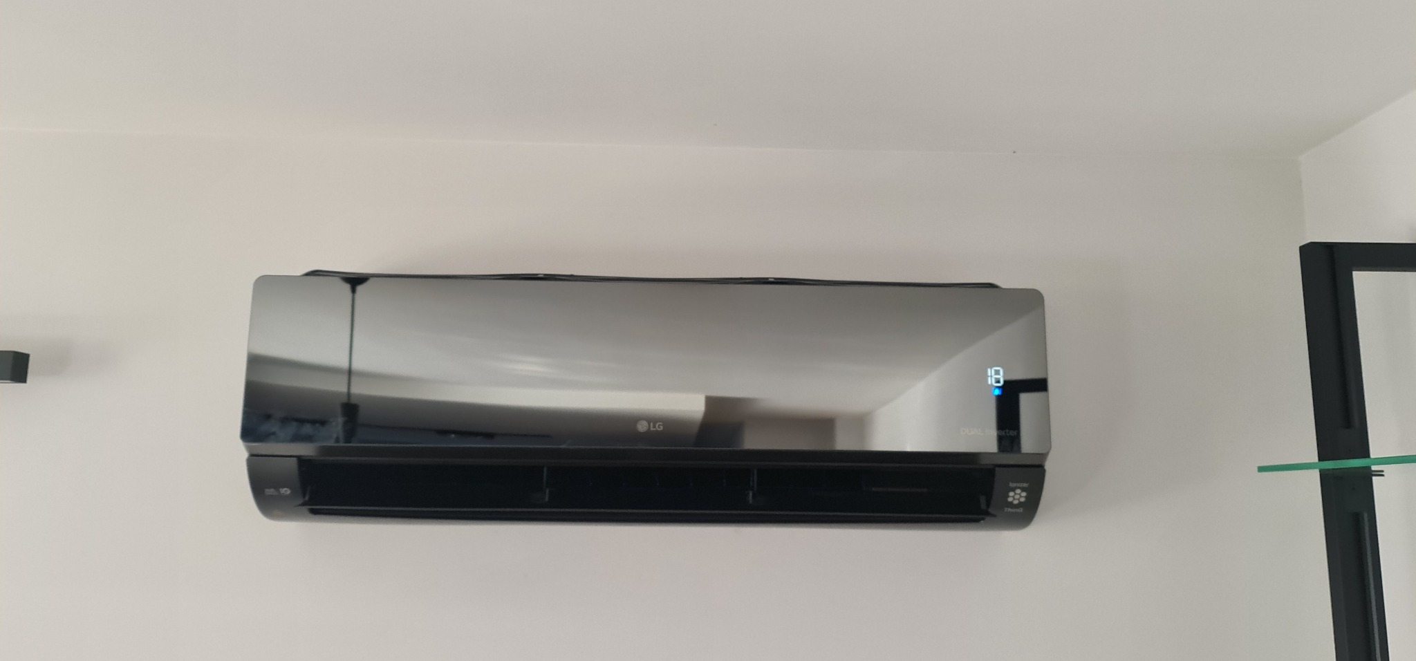 Czarny klimatyzator LG Dual Inverter zamontowany na białej ścianie, wyświetlacz pokazuje 18 stopni, widoczne odbicie wnętrza pomieszczenia.