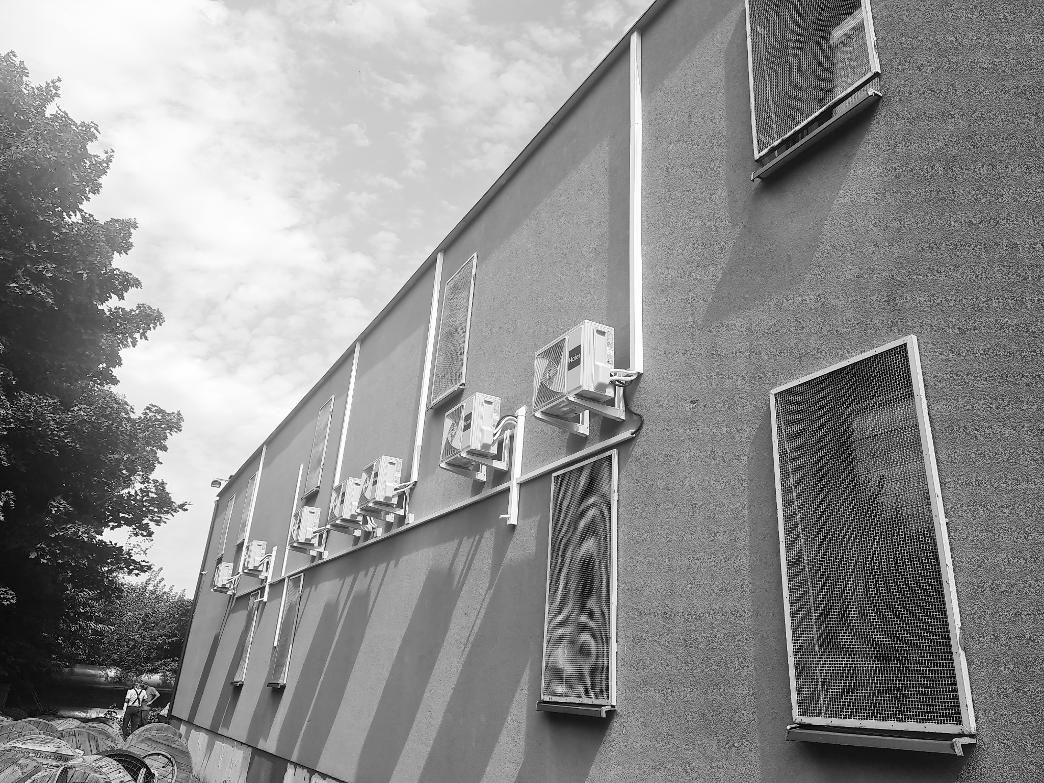 Czarno-białe zdjęcie ściany budynku z zainstalowanymi klimatyzatorami Haier i oknami zabezpieczonymi siatką, widoczne również zwinięte kable na szpulach i zieleń drzew