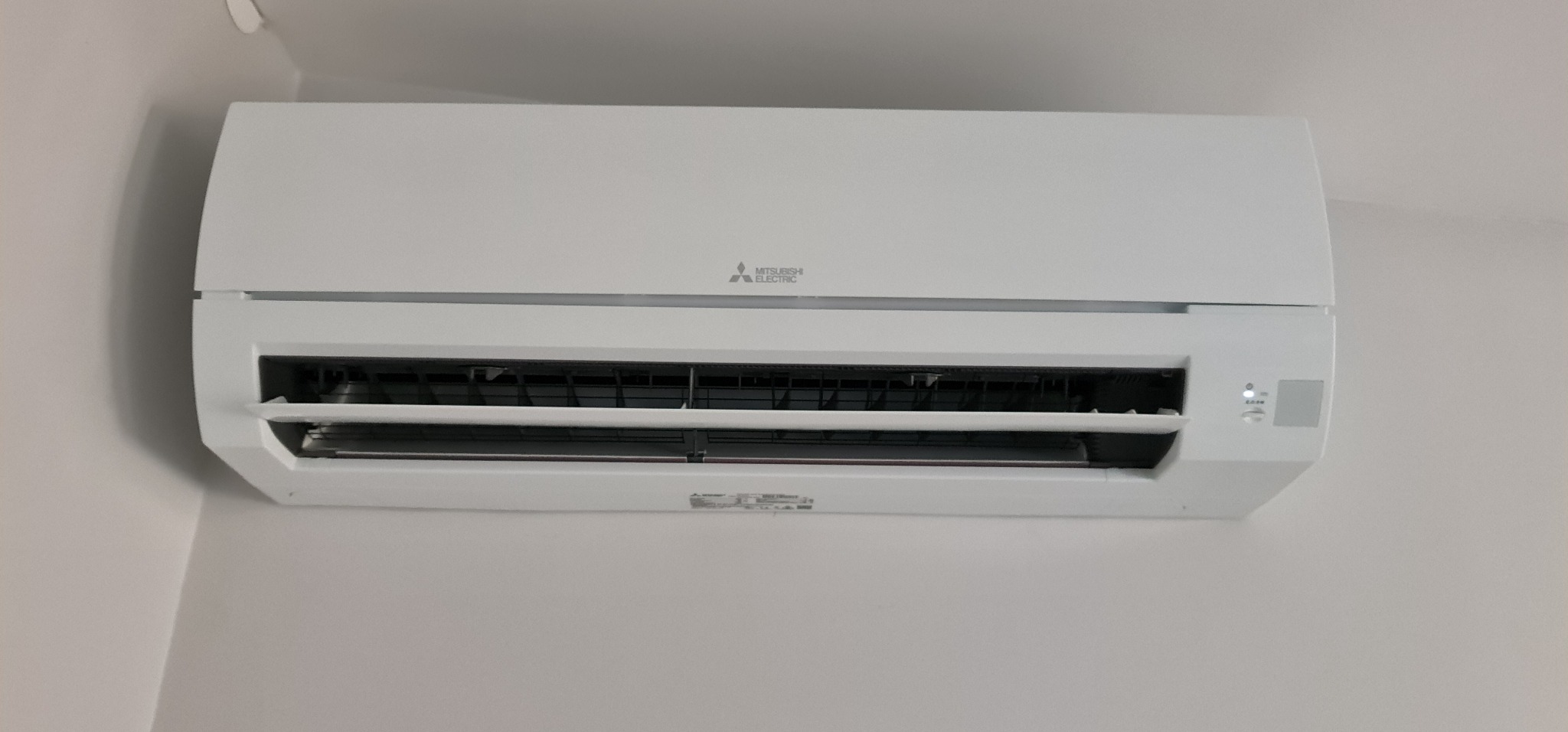Biała jednostka wewnętrzna klimatyzacji Mitsubishi Electric zamontowana na ścianie, widoczna dioda sygnalizacyjna stanu pracy.