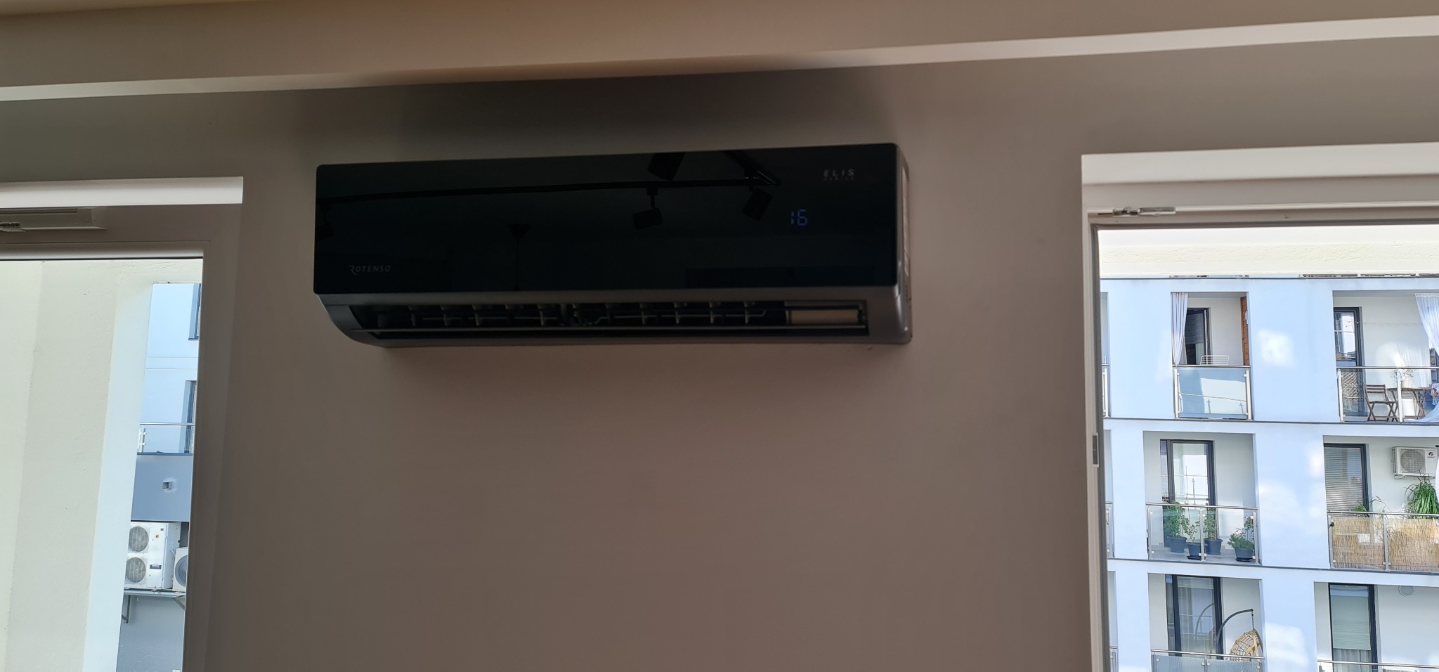 Czarny klimatyzator marki Rotenso Elis zamontowany na ścianie w jasnym pomieszczeniu, widoczny fragment okna z widokiem na balkon innego budynku.