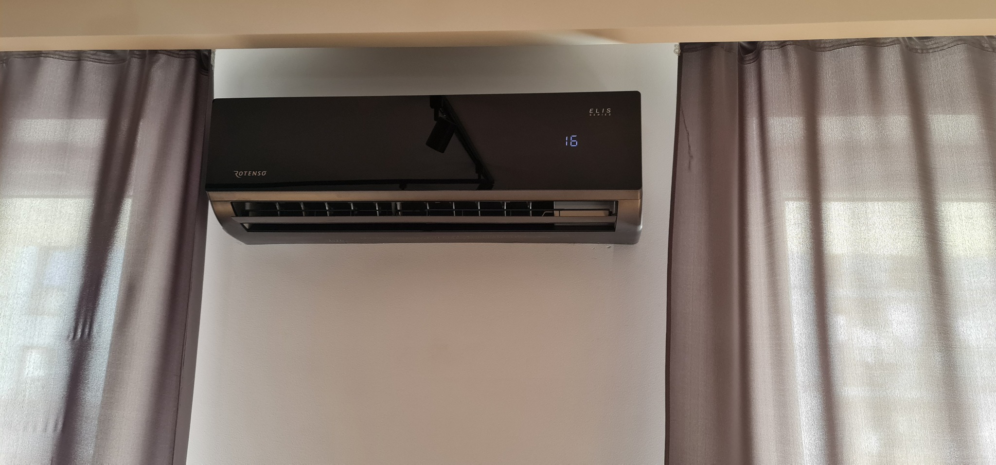 Czarny klimatyzator Rotenso Elis Series zamontowany na białej ścianie, widoczne szare zasłony po bokach.