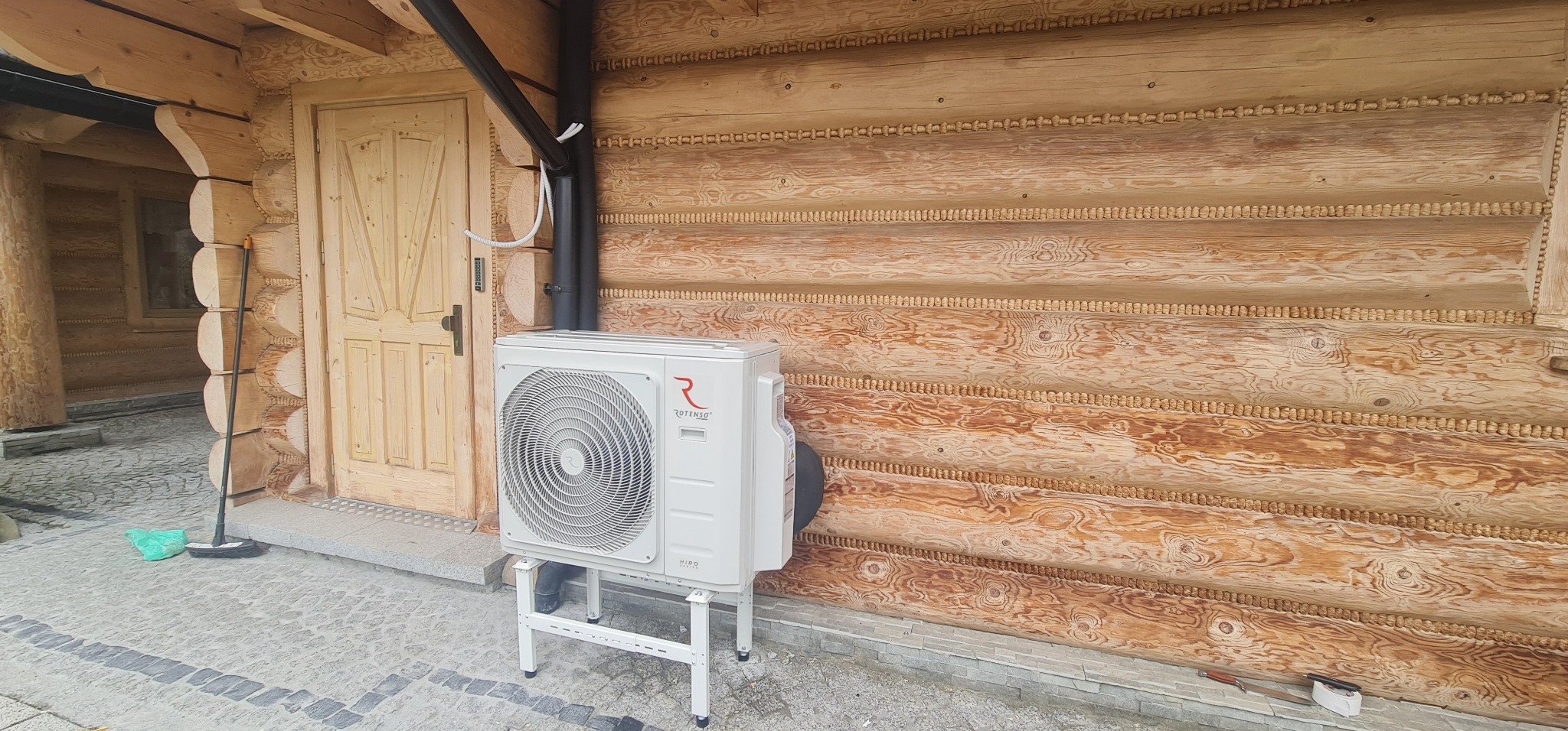 Biała jednostka zewnętrzna klimatyzacji marki Rotenso zamontowana na metalowym stelażu przed drewnianym domem z bali, widoczne rury odprowadzające.