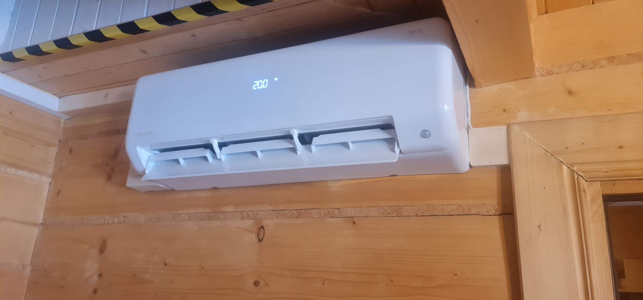 Biały klimatyzator zamontowany na drewnianej ścianie wewnątrz pomieszczenia, wyświetlający temperaturę 20 stopni Celsjusza.