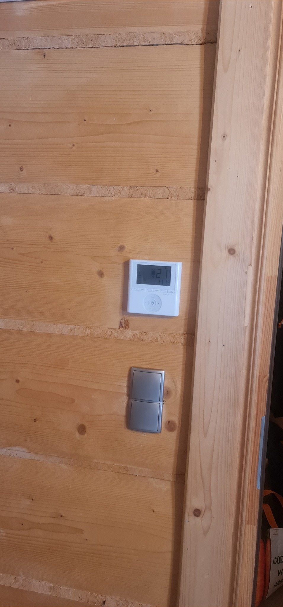 Biały panel sterowania klimatyzacją z wyświetlaczem temperatury 21 stopni Celsjusza, zamontowany na drewnianej ścianie obok podwójnego włącznika światła.