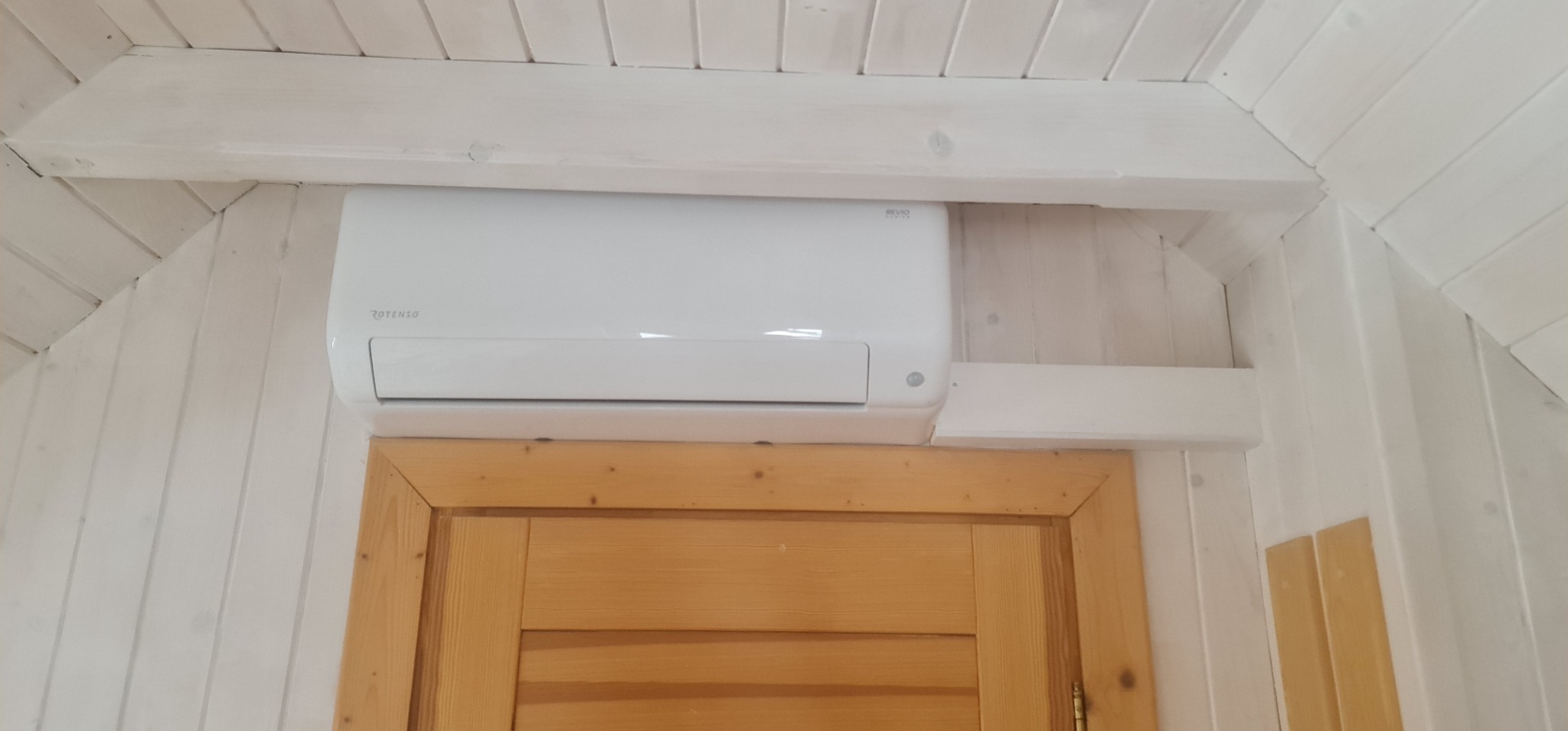 Biała jednostka klimatyzacyjna Rotenso zamontowana w drewnianym domu z boazerią, widoczna nad futryną drzwi.