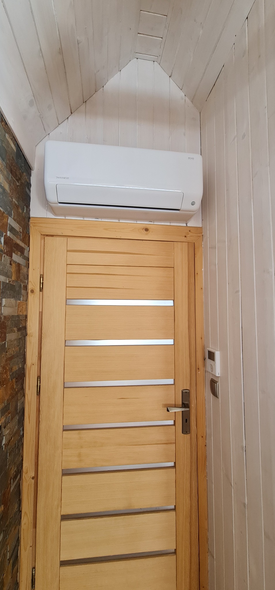 Biały klimatyzator zamontowany nad drewnianymi drzwiami z poziomymi, metalowymi wstawkami w pomieszczeniu wykończonym boazerią i kamieniem dekoracyjnym.