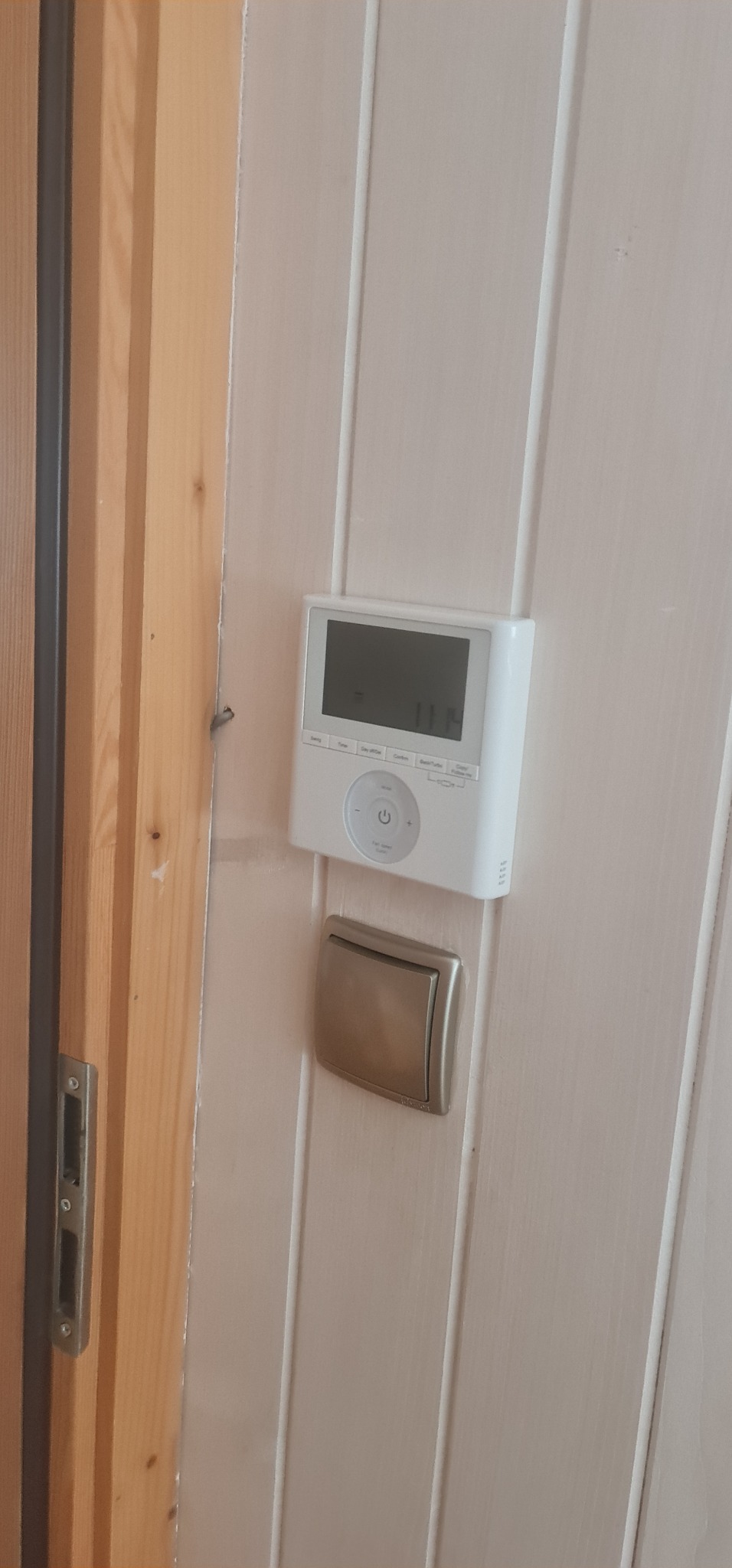 Biały panel sterowania klimatyzacją z wyświetlaczem LCD i przyciskiem włącznika zamontowany na panelowej ścianie obok drewnianej futryny drzwi.