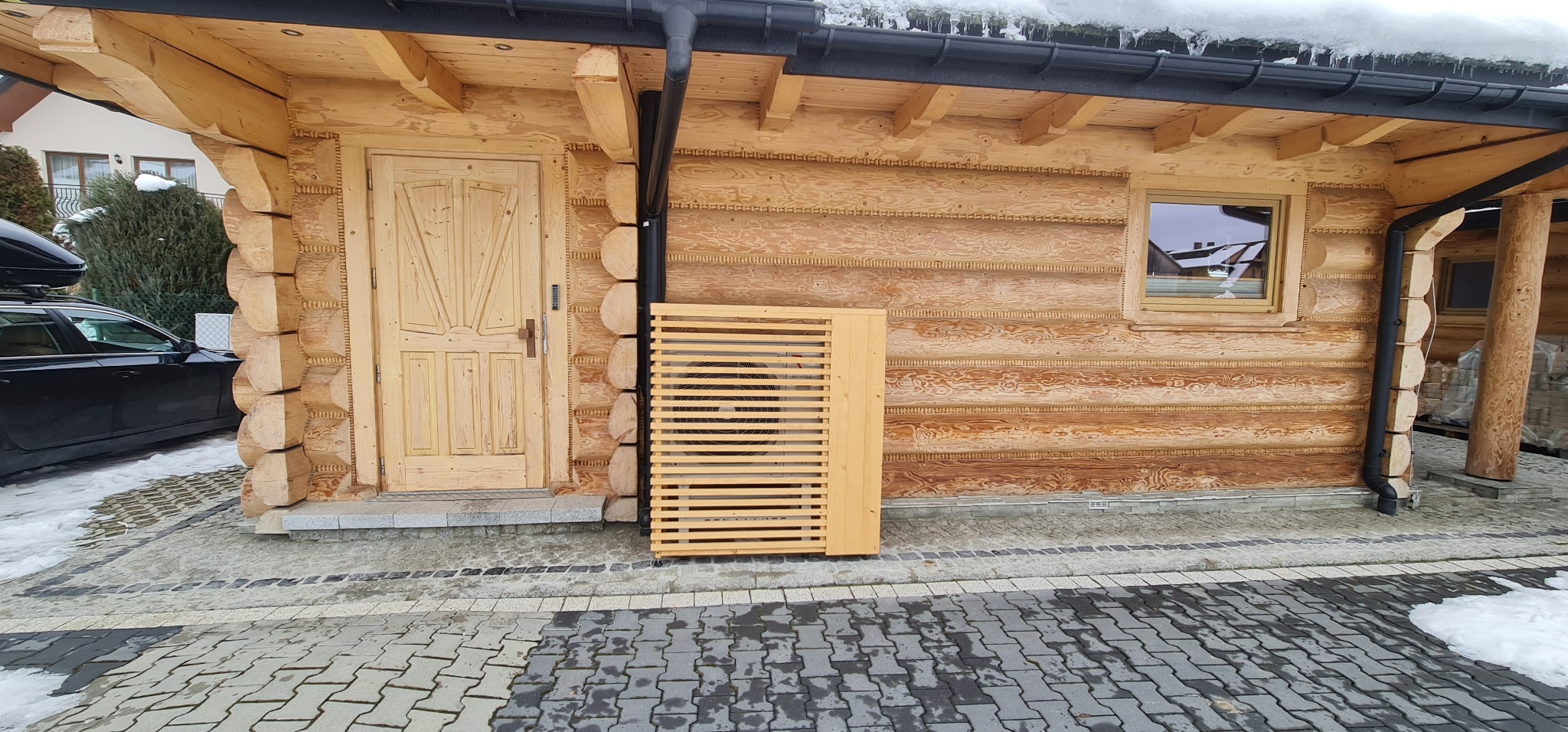 Drewniana obudowa jednostki zewnętrznej klimatyzatora przy ścianie domu z bali, widoczna czarna rynna i fragment kostki brukowej.