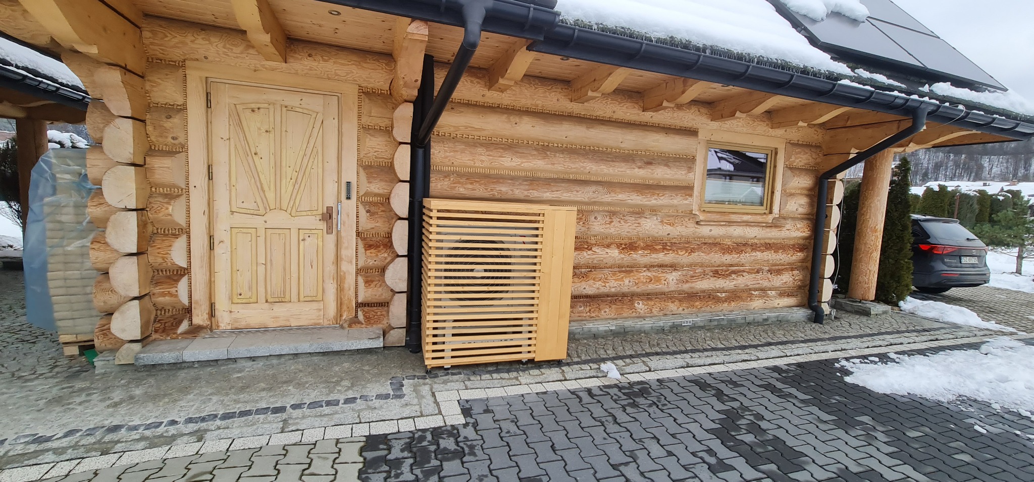 Zewnętrzna jednostka klimatyzacji zamontowana przy drewnianym domu, osłonięta drewnianą obudową z poziomych listew, widoczna elewacja z bali i fragment podjazdu z kostki brukowej.