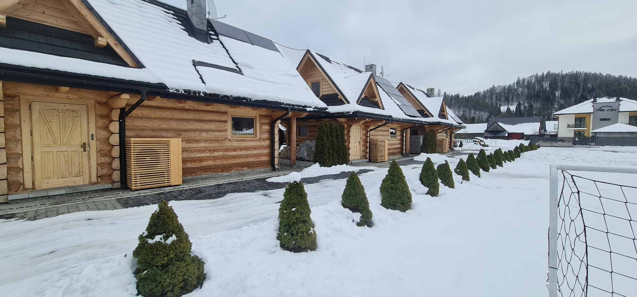 Zewnętrzne jednostki klimatyzacyjne w drewnianych obudowach przy domkach w stylu góralskim, zimowy krajobraz z ośnieżonymi drzewami.