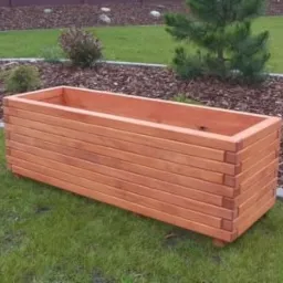 Prostokątna, drewniana donica ogrodowa z poziomych desek, umieszczona na trawniku z korą drzewną w tle.