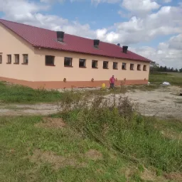 Budynek gospodarczy z nową elewacją w odcieniach beżu i brązu, czerwony dach z kominami wentylacyjnymi, trawa i nieutwardzony teren wokół, pracownik w różowej koszulce w tle.