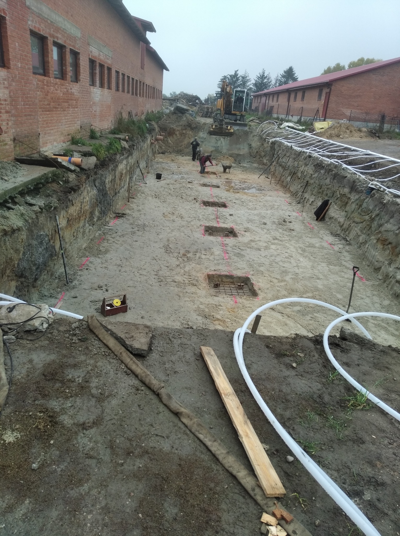 Budowa fundamentów pod budynek murowany: wykop, zbrojenie, rury instalacyjne i pracownicy w tle.