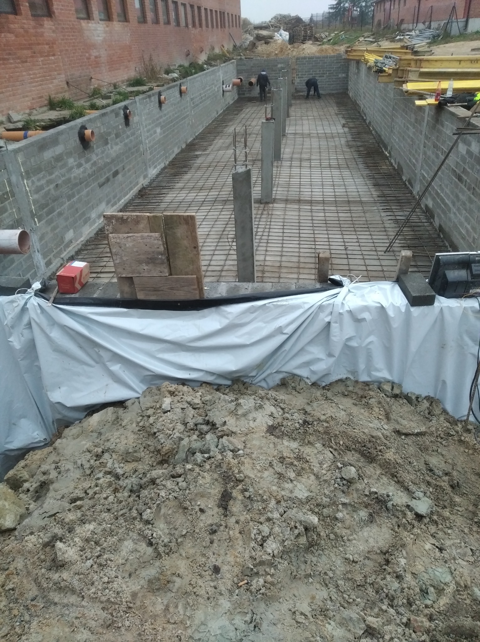 Budowa fundamentów domu w Milanowie: widok na wykop z ułożonym zbrojeniem i wylanymi słupami, zabezpieczony folią i obsypany ziemią.