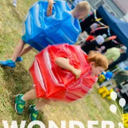 Dwóch chłopców w dmuchanych kostiumach sumo, jeden czerwony i jeden niebieski, biegnie po trawie podczas imprezy plenerowej.