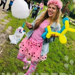 Uśmiechnięta dziewczyna w różowej tiulowej spódnicy i kwiecistym wianku trzyma balonowego pieska w barwach niebiesko-żółtych oraz watę cukrową, w tle grupa dzieci i zielona trawa.