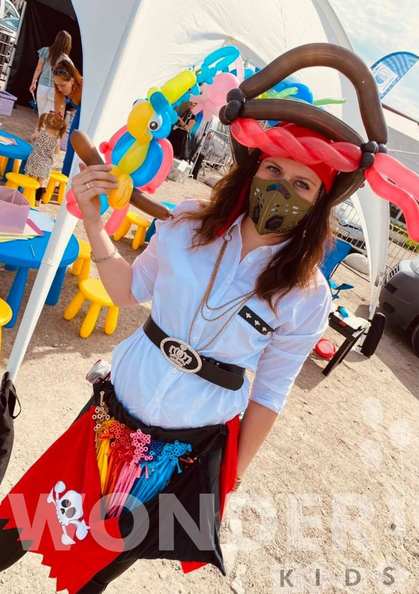 Kobieta w przebraniu pirata z balonowym papugą i kapeluszem z balonów, pozująca na tle kolorowych stolików i dmuchanego namiotu podczas imprezy plenerowej.