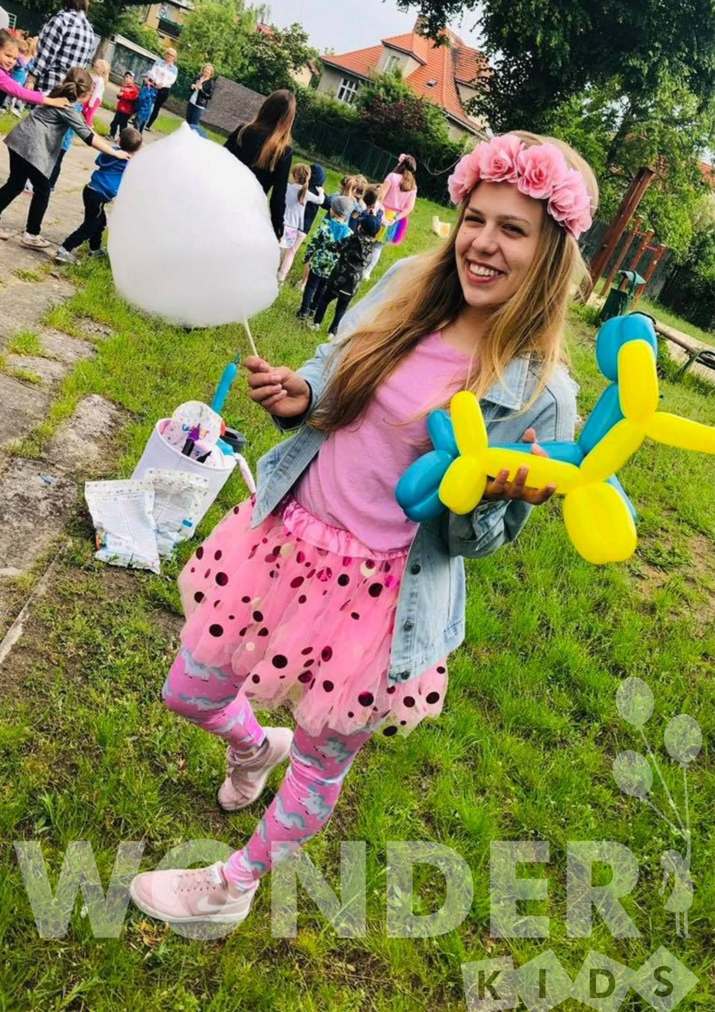 Uśmiechnięta dziewczyna w różowej tiulowej spódnicy i kwiecistym wianku trzyma balonowego pieska w barwach niebiesko-żółtych oraz watę cukrową, w tle grupa dzieci i zielona trawa.