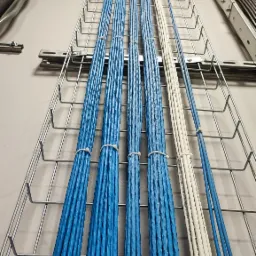 Równoległe wiązki niebieskich i białych kabli UTP ułożone w metalowej korycie kablowej, przymocowane opaskami zaciskowymi, widoczne fragmenty etykiet na kablach.