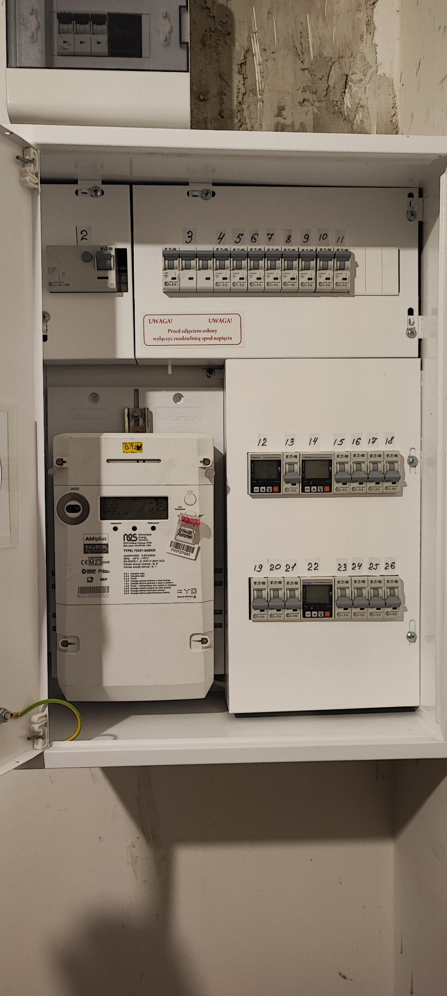 Otwarta szafka elektryczna z licznikiem i zabezpieczeniami, widoczne numery obwodów, naklejki ostrzegawcze i przewód uziemiający.