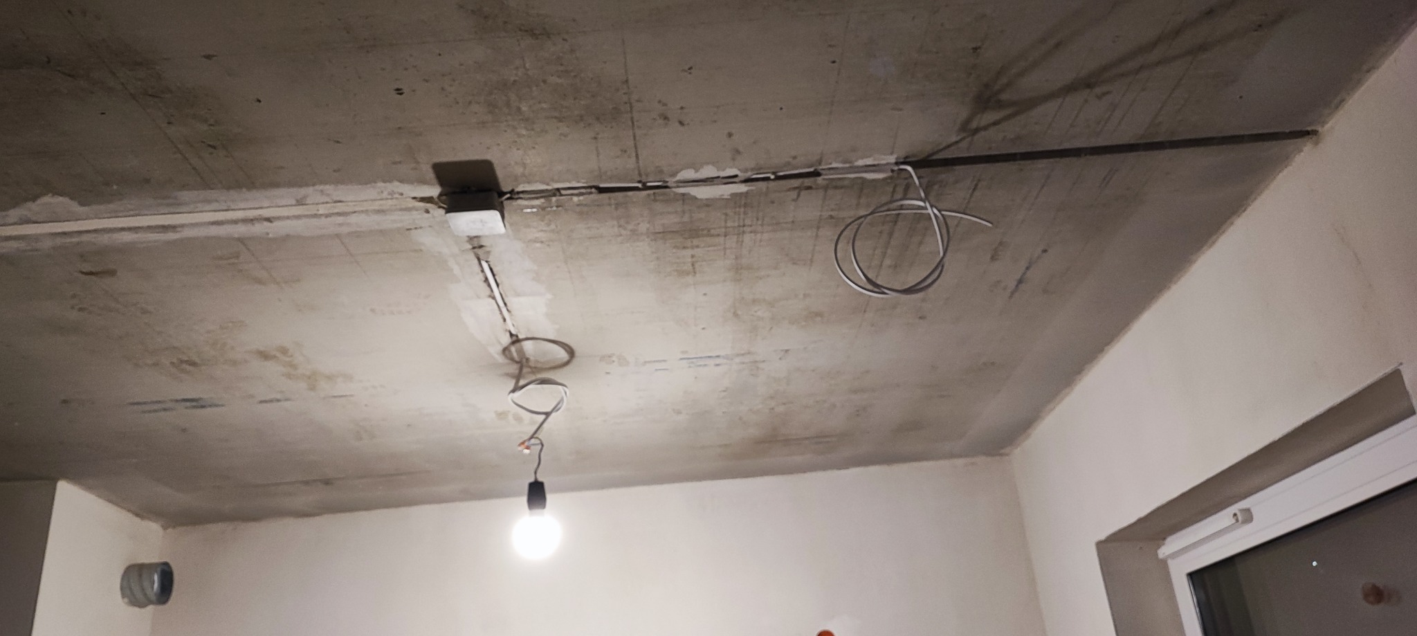 Surowy betonowy sufit z widoczną instalacją elektryczną: przewody, puszka i prowizoryczna żarówka.