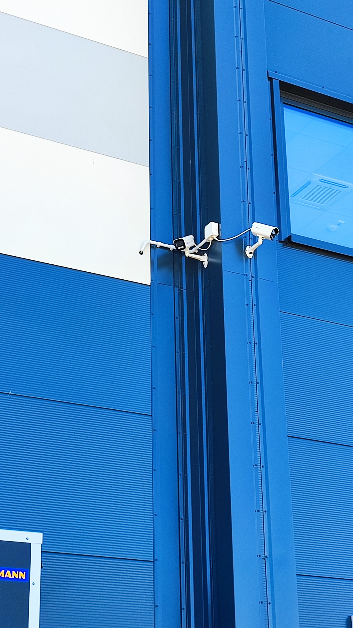 Zewnętrzny montaż trzech kamer monitoringu na niebieskiej, metalowej elewacji budynku z widocznym oknem i fragmentem białej ściany.