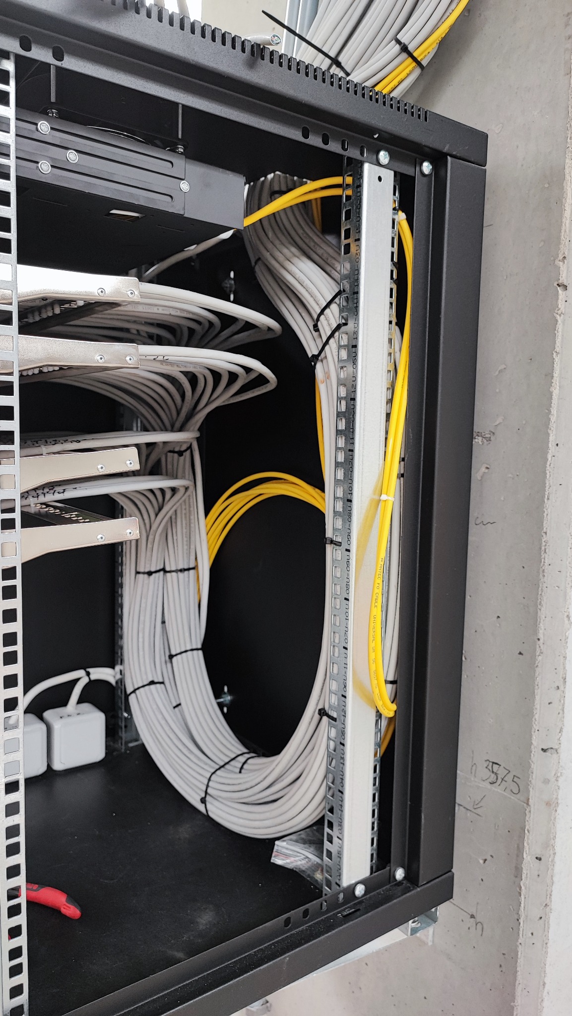 Kable sieciowe i światłowodowe ułożone w szafie rack, przewody białe i żółte, uporządkowane za pomocą opasek zaciskowych, widoczna listwa montażowa i fragment ściany z betonową fakturą.