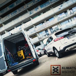 Klimatyzacja Wschód Mobilny Serwis Klimatyzacji - Mobilny serwis. Otwarty van z narzędziami do klimatyzacji samochodowej na tle białego auta osobowego i budynku mieszkalnego. Widoczny sprzęt R-1234 MP.