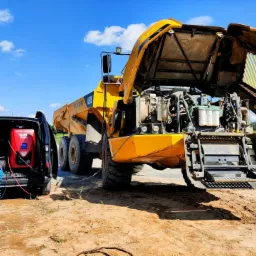 Żółta wywrotka Volvo A30E z otwartą maską silnika, obok czarny bus z czerwonym urządzeniem diagnostycznym i przewodami, na piaszczystym placu w słoneczny dzień.