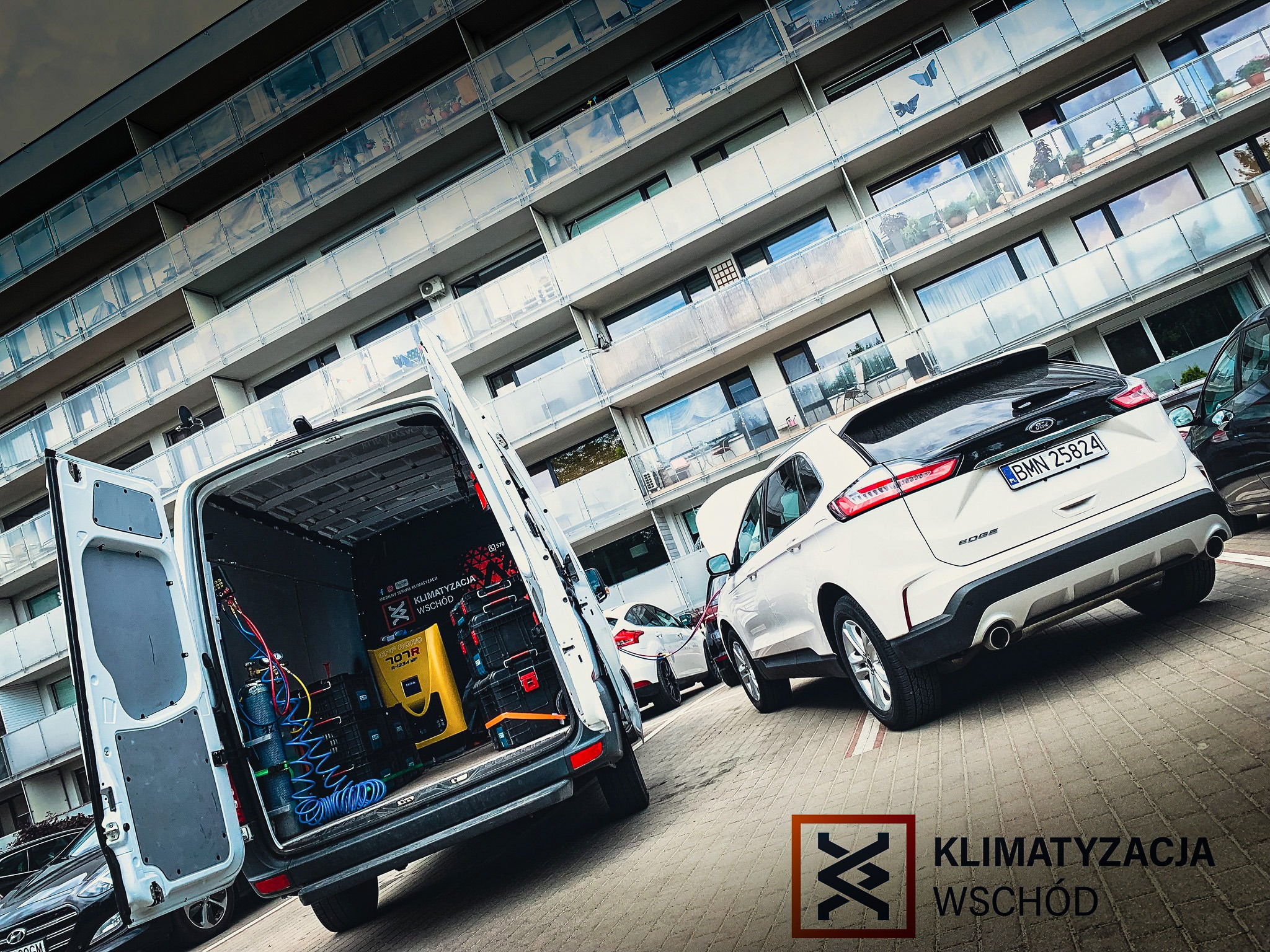 Mobilny serwis. Otwarty van z narzędziami do klimatyzacji samochodowej na tle białego auta osobowego i budynku mieszkalnego. Widoczny sprzęt R-1234 MP.