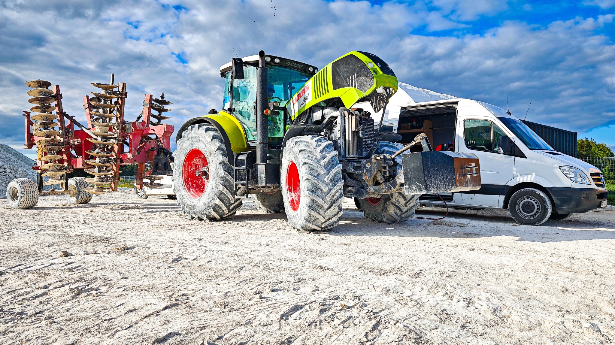 Zielono-czarny traktor Claas Axion z podniesioną maską, podpięty do czerwonego agregatu talerzowego, stoi na placu obok białego busa serwisowego.