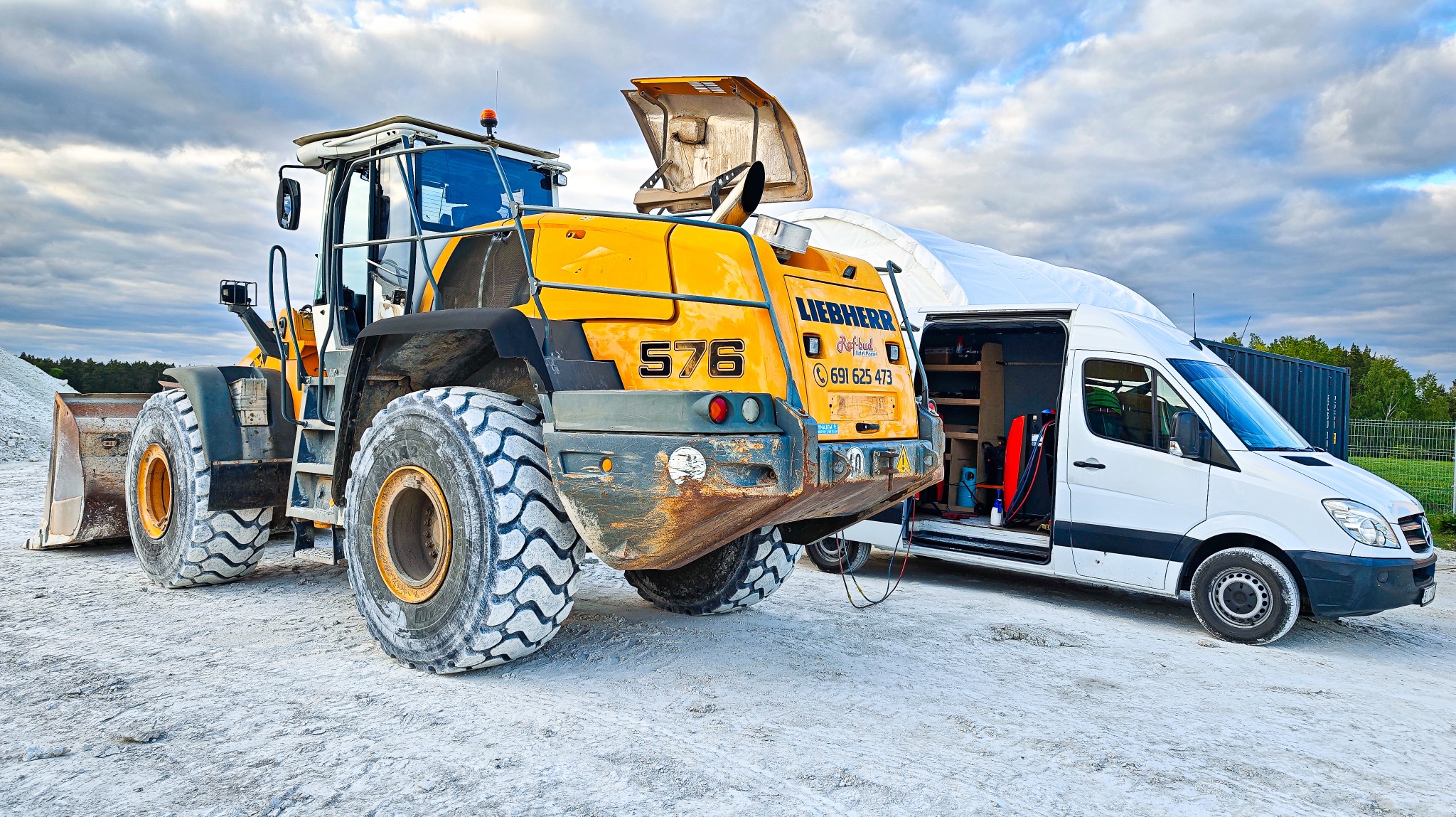 Żółta ładowarka Liebherr 576 z otwartą maską silnika, obok biały van z otwartymi drzwiami, na placu budowy pod pochmurnym niebem.