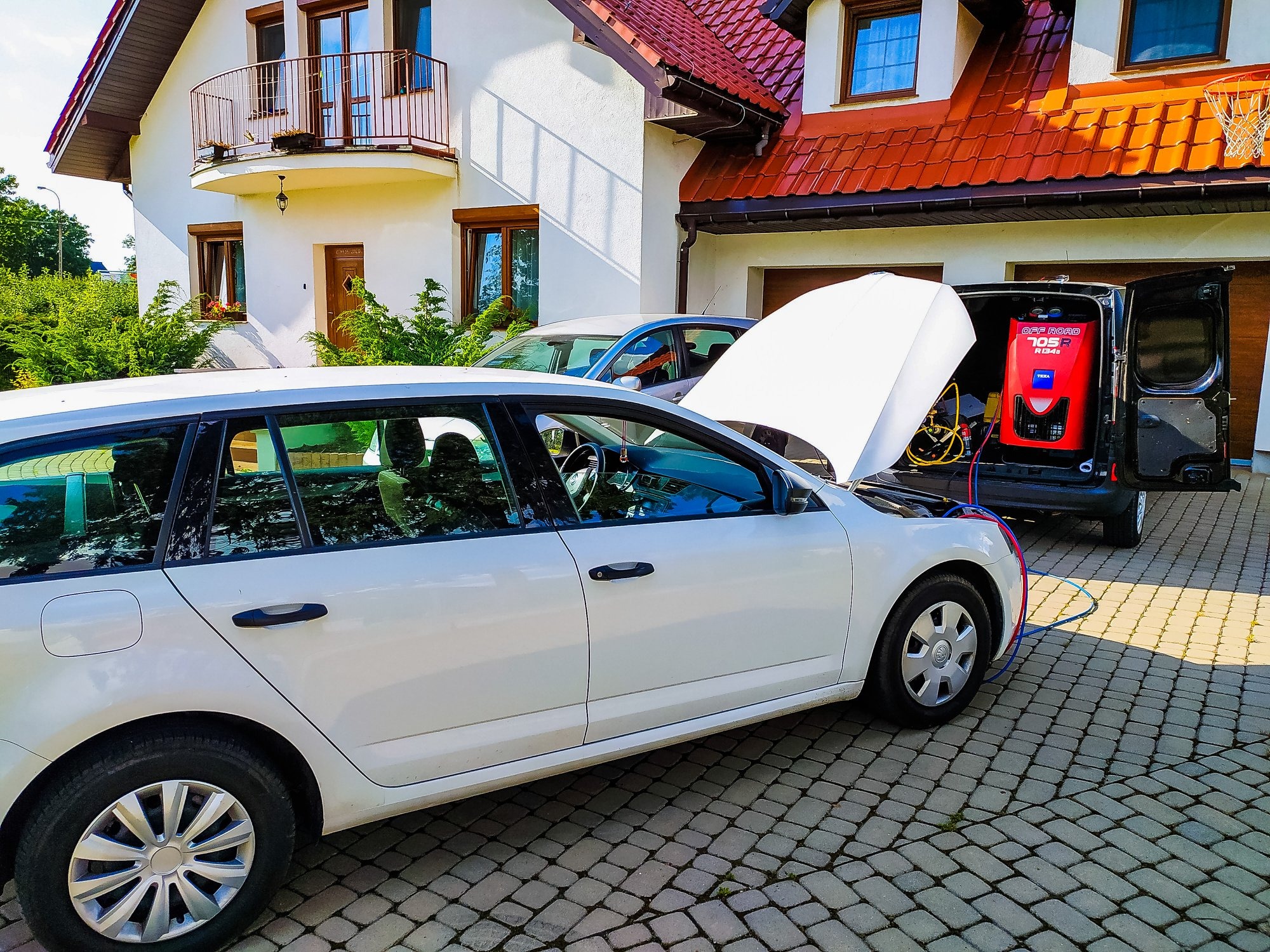 Biały samochód kombi z otwartą maską, podłączony do czerwonego urządzenia diagnostycznego w bagażniku czarnego vana na tle domu z czerwonym dachem i brukowanym podjazdem.