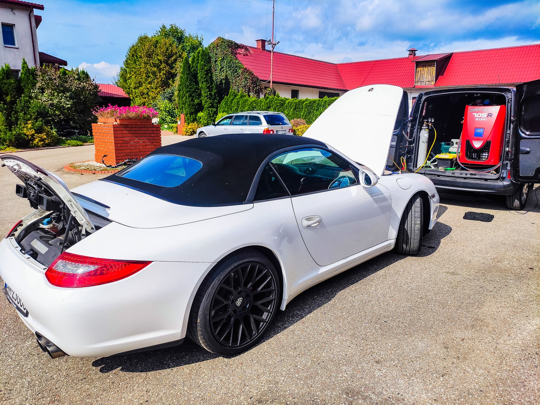 Biały kabriolet Porsche z otwartą maską i bagażnikiem, podłączony do urządzenia do serwisu klimatyzacji w zaparkowanym vanie na zewnątrz, widoczne budynki mieszkalne w tle.