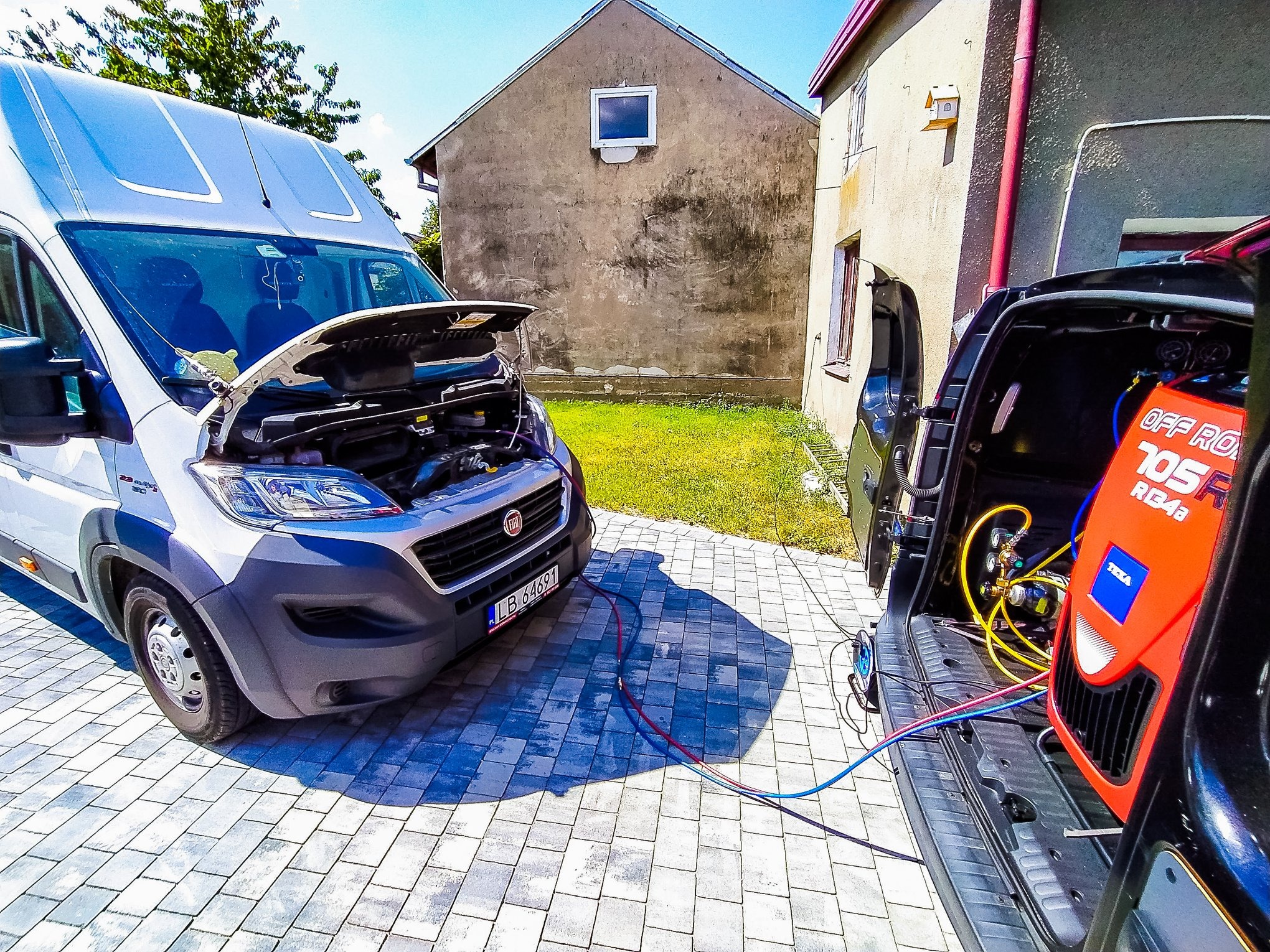 Mobilny serwis klimatyzacji samochodowej: biały van Fiat z otwartą maską i podłączonym urządzeniem do napełniania klimatyzacji, stojący na kostce brukowej przed budynkiem.