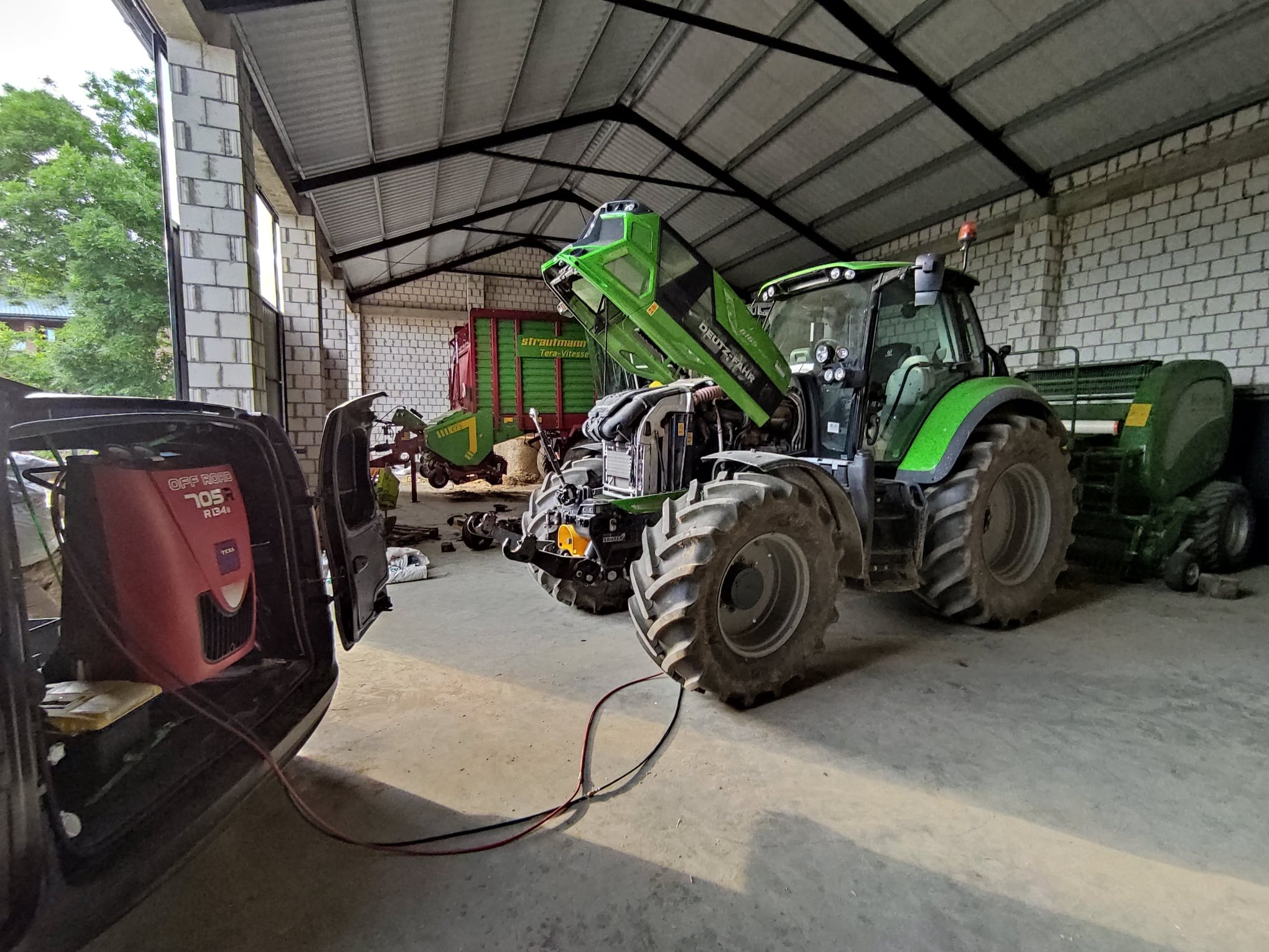 Zielony traktor Deutz-Fahr z podniesioną maską wewnątrz budynku, podłączony do czerwonego urządzenia diagnostycznego Texa Off Road 705R R134a, sugerującego serwis lub naprawę.