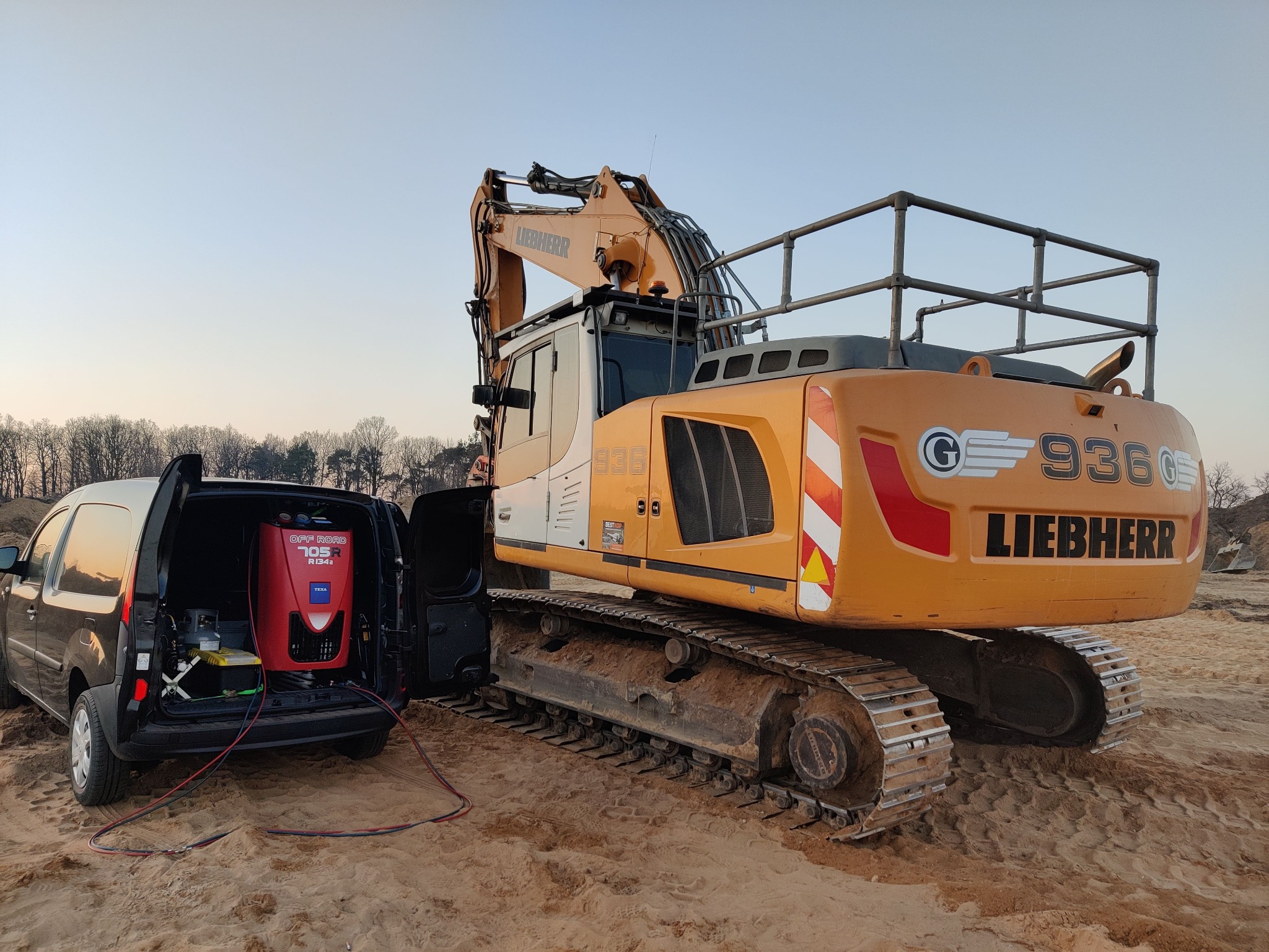 Serwis klimatyzacji mobilnej przy pomarańczowej koparce Liebherr 936 na piaszczystym terenie, z otwartym bagażnikiem czarnego vana i widocznym urządzeniem Texa Off Road 705R R134a.