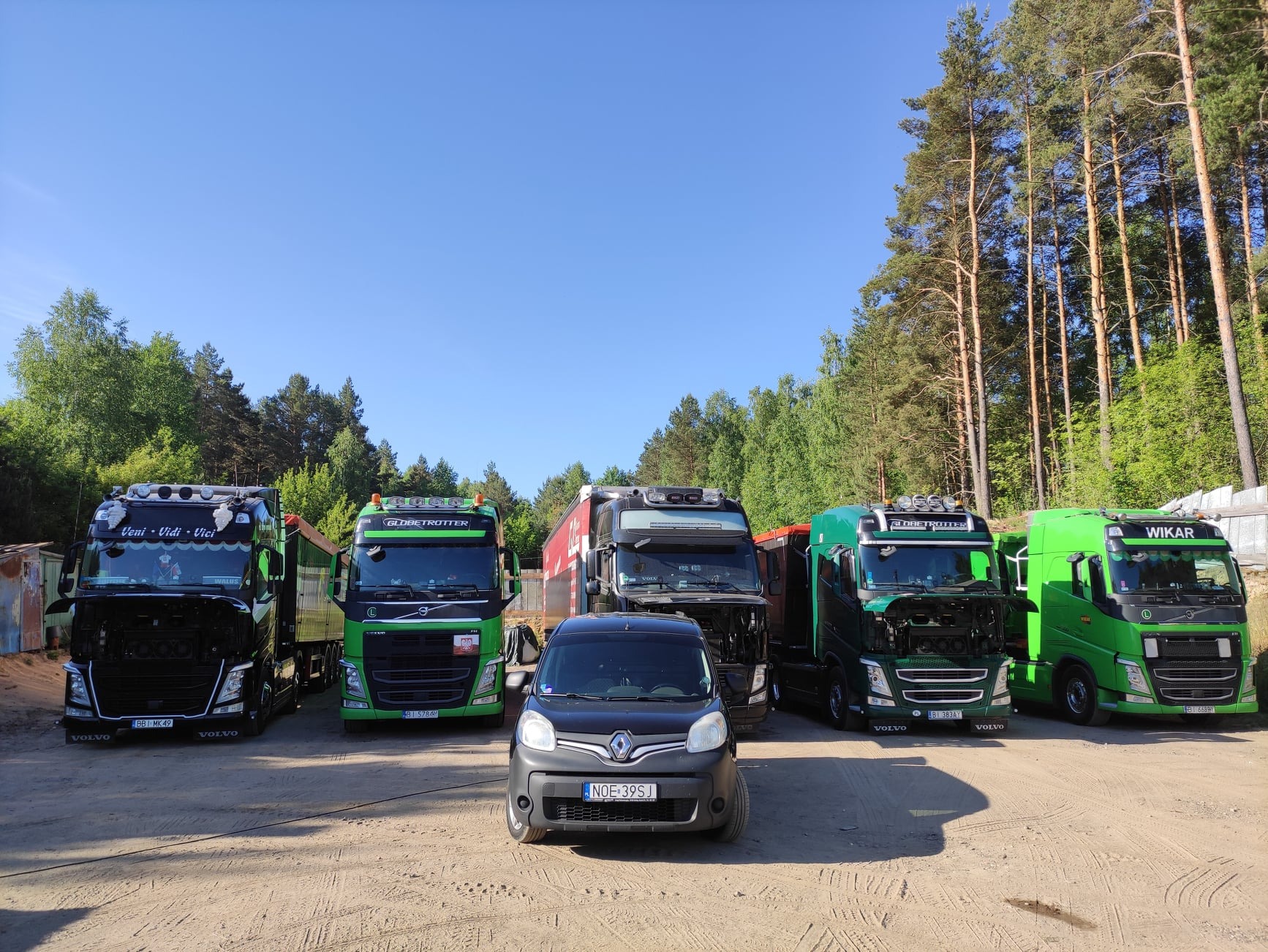Flota ciężarówek Volvo różnych modeli i kolorów, zaparkowanych na piaszczystym placu w słoneczny dzień, z małym czarnym samochodem osobowym na pierwszym planie.