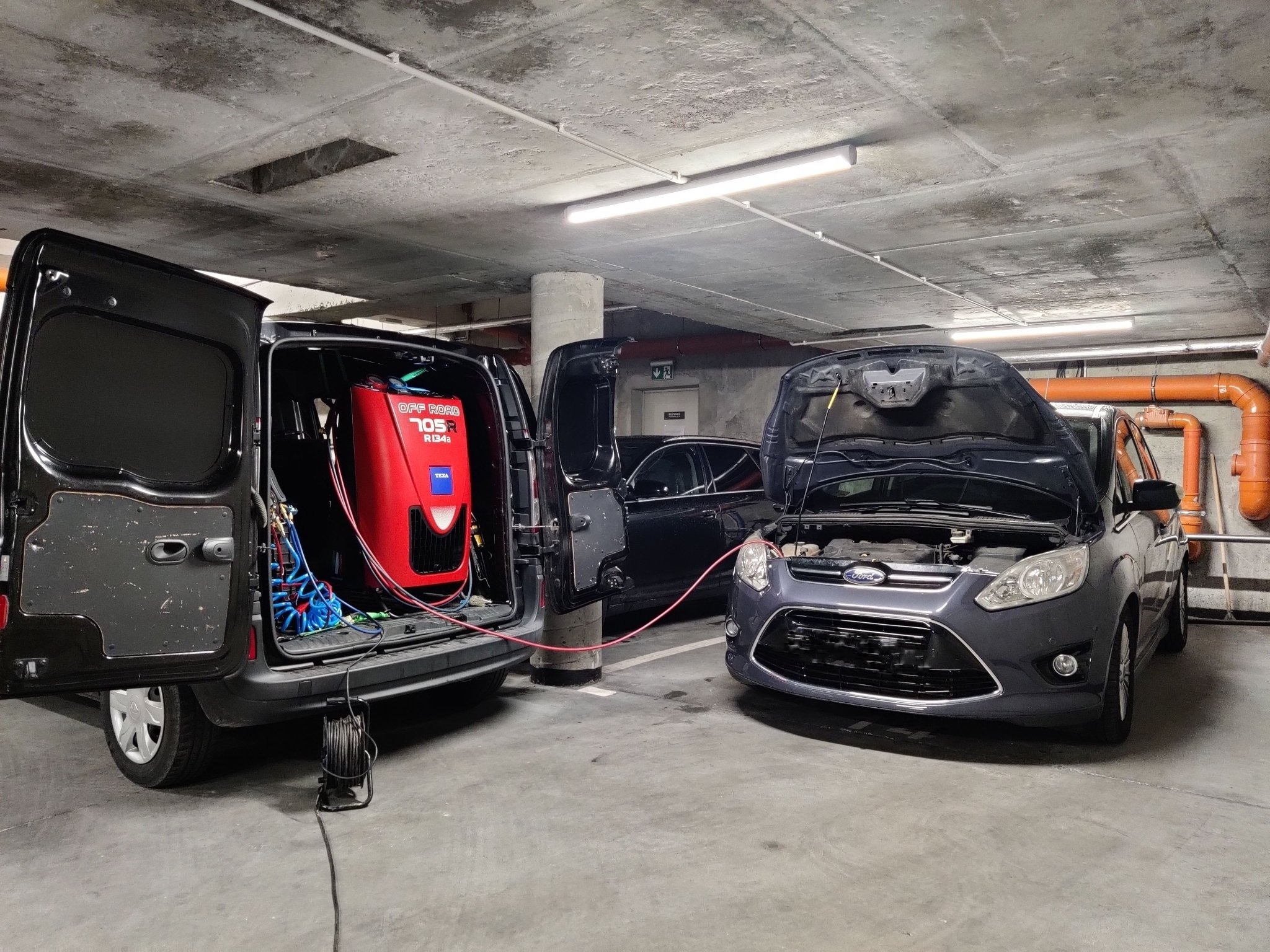 Szary Ford z otwartą maską podłączony przewodem do czerwonego urządzenia w bagażniku czarnego vana na parkingu podziemnym.