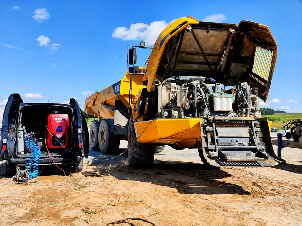 Żółta wywrotka Volvo A30E z otwartą maską silnika, obok czarny bus z czerwonym urządzeniem diagnostycznym i przewodami, na piaszczystym placu w słoneczny dzień.