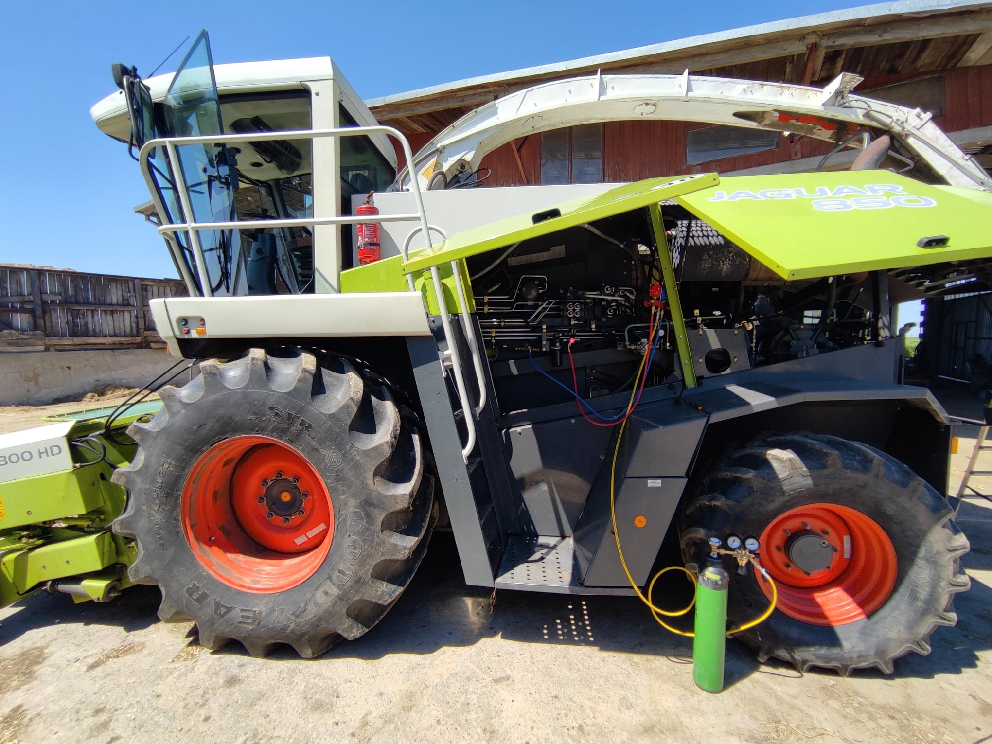 Zielony kombajn rolniczy Jaguar 850 z otwartą maską silnika, podłączony do butli z gazem, prawdopodobnie podczas serwisu lub naprawy.