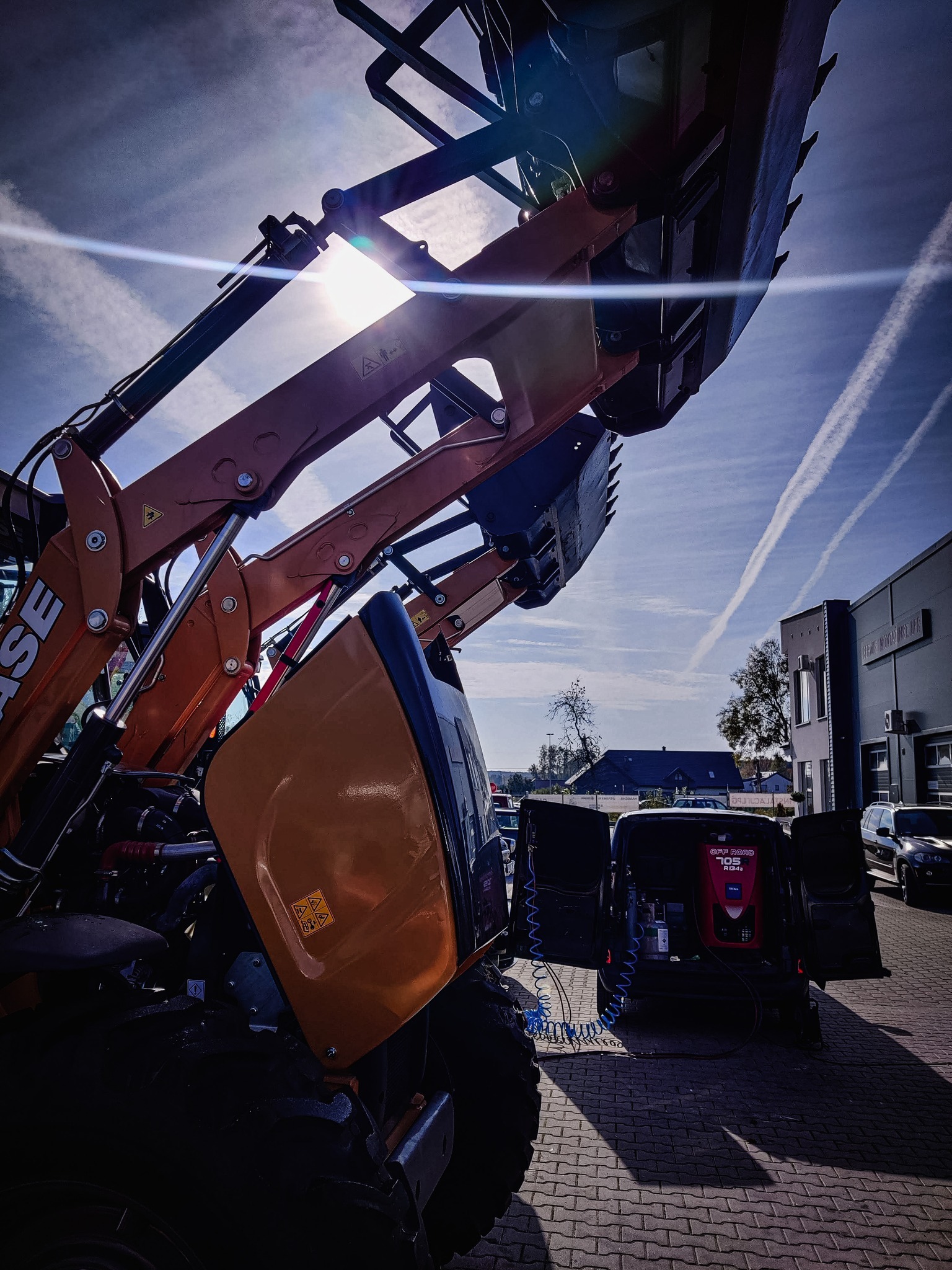 Pomarańczowa ładowarka teleskopowa CASE z podniesioną łyżką na tle błękitnego nieba z smugami kondensacyjnymi, obok zaparkowany czarny van z otwartymi drzwiami i urządzeniem do obsługi klimatyzacji.