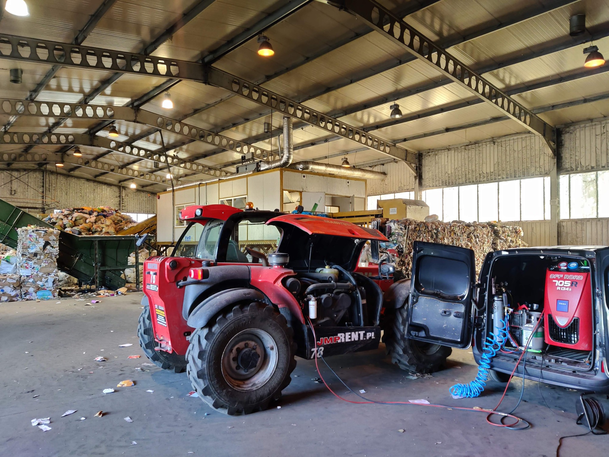 Czerwony wózek widłowy z otwartą maską silnika i podłączonymi przewodami, obok zaparkowany samochód z urządzeniem do klimatyzacji samochodowej w otwartym bagażniku, w tle hala z belami makulatury.