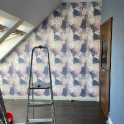 Ściana w pokoju na poddaszu po tapetowaniu w geometryczny wzór, widoczna drabina aluminiowa i fragment listwy przypodłogowej.