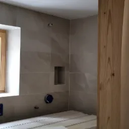 Łazienka w trakcie wykończenia: szare płytki ceramiczne na ścianach, drewniana rama okienna z widokiem na zewnątrz, podłoga przygotowana pod ułożenie płytek, widoczne elementy instalacji podtynkowych.